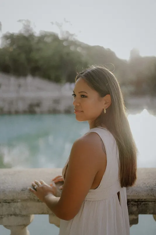 Woman in a white dress gazes softly over a stone railing by the water during a couples & engagements session.