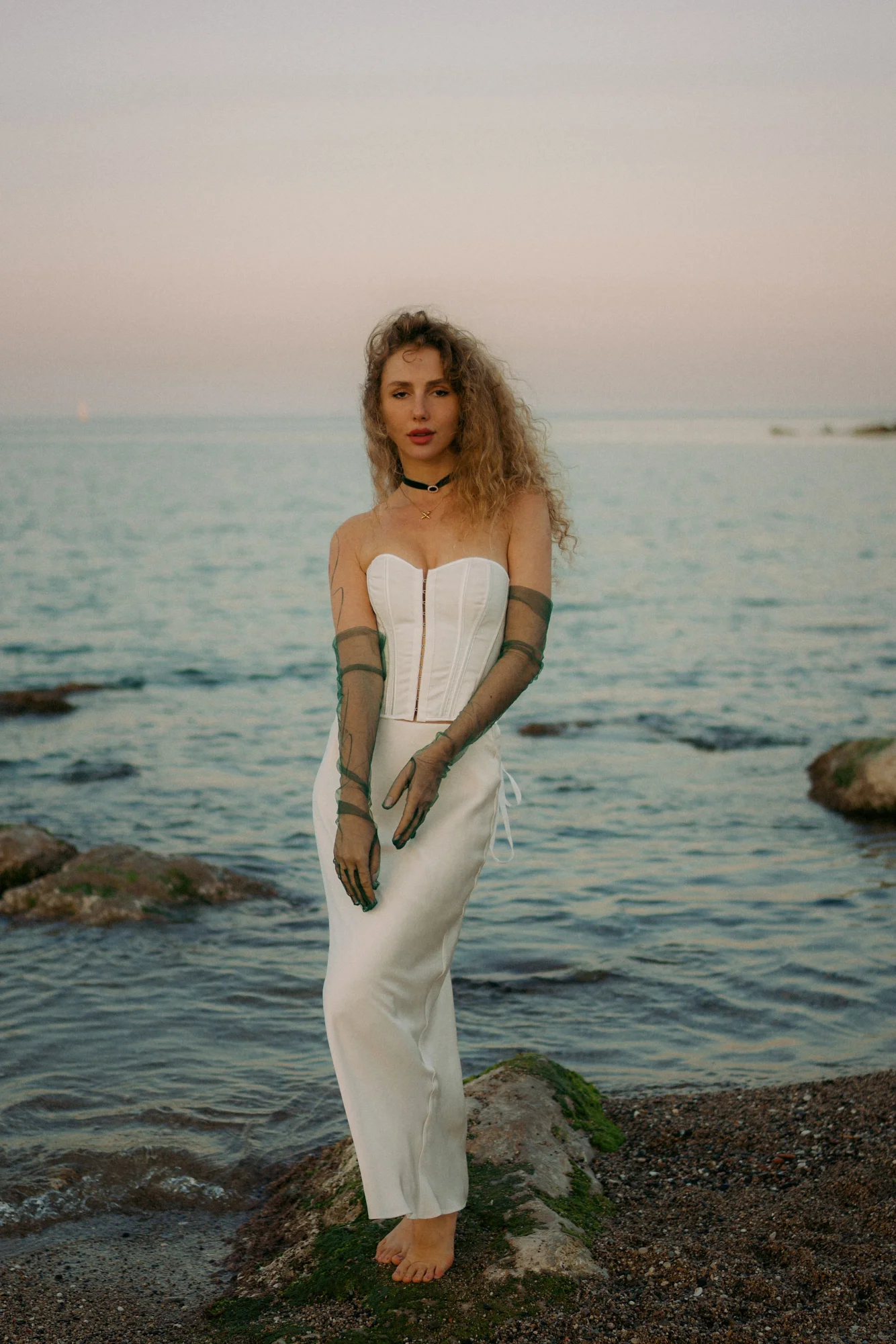 Woman in a white dress poses barefoot on seaside rocks at sunset during a Vacation Portraits photoshoot.