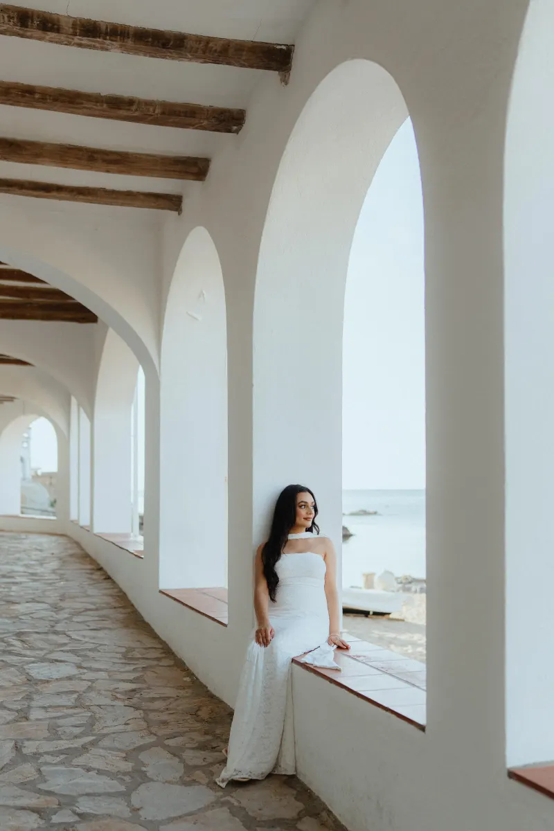 Woman in a white dress sits by a seaside archway, smiling softly during a surprise proposal photoshoot.