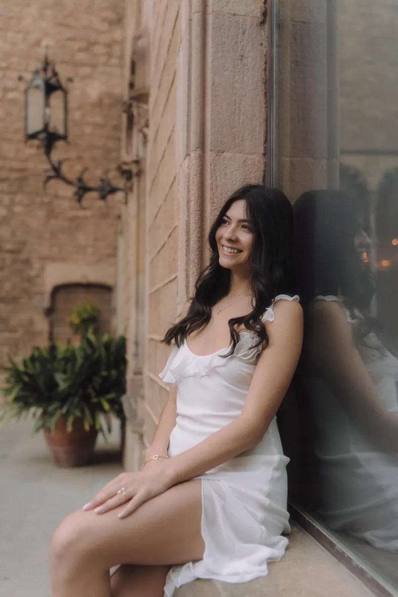 Woman in a white dress sits by a stone wall, smiling softly during a couples & engagements photoshoot in a historic courtyard