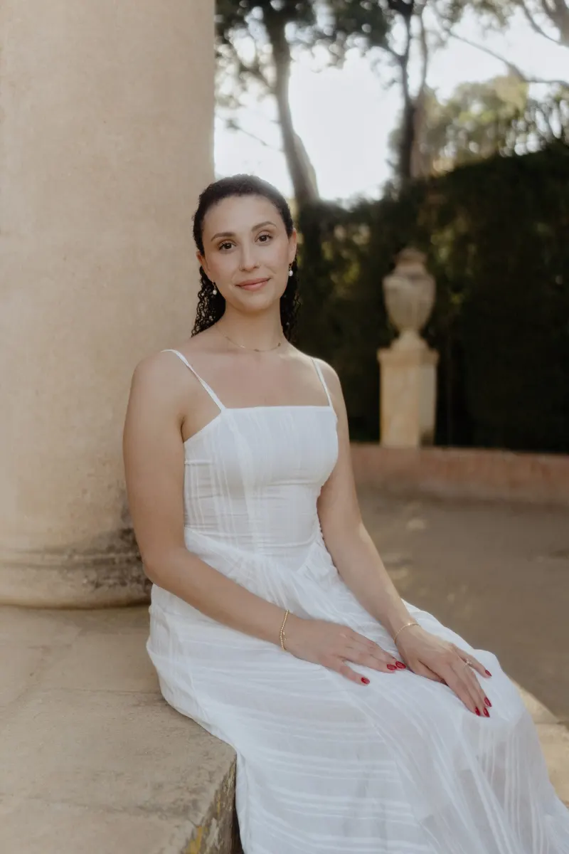 Woman in a white dress sits gracefully by a stone column, softly smiling during a couples & engagements photoshoot.