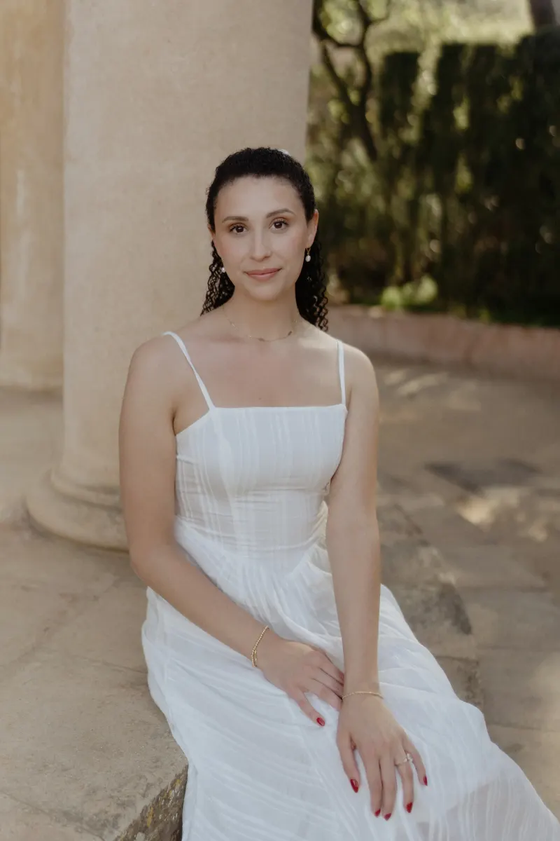 Woman in a white dress sits gracefully by a stone column, softly smiling during a couples & engagements session.