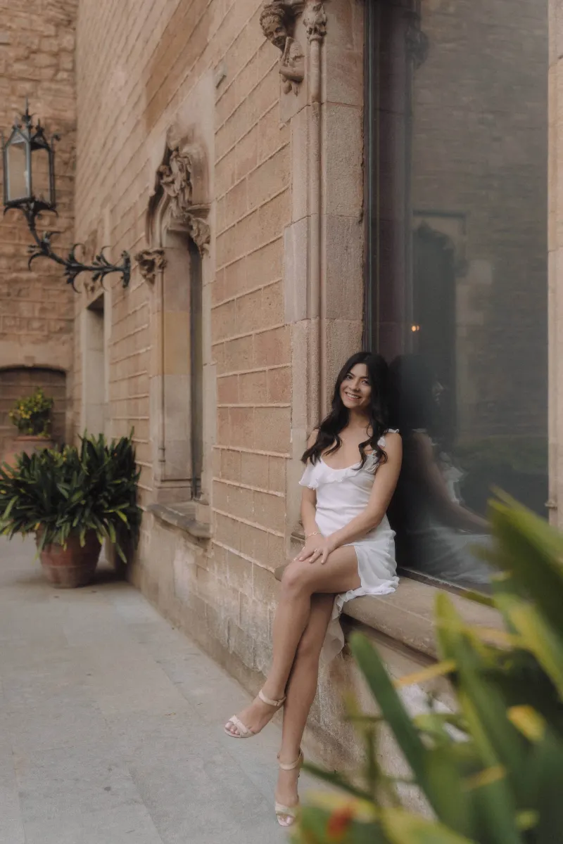 Woman in a white dress sits on a stone window ledge, smiling softly during a couples & engagements photoshoot.