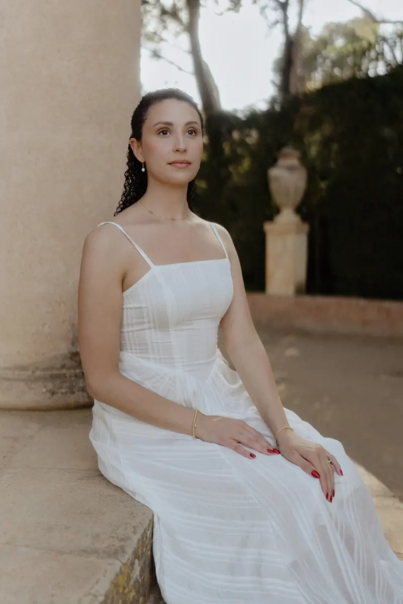 Woman in a white dress sits serenely by a stone column, captured during a couples & engagements photoshoot in a garden.