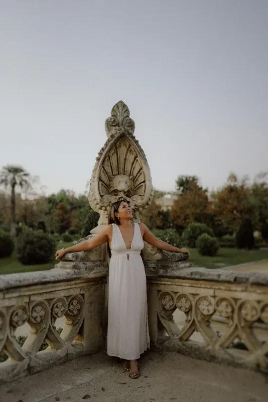 Woman in a white dress smiles and leans back on ornate stone railing during a couples photoshoot in a historic garden.