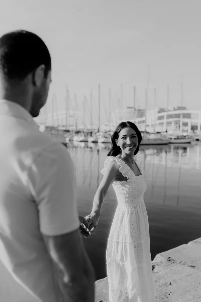 Woman in a white dress smiles at her partner, holding hands by the marina during a couples & engagements session.