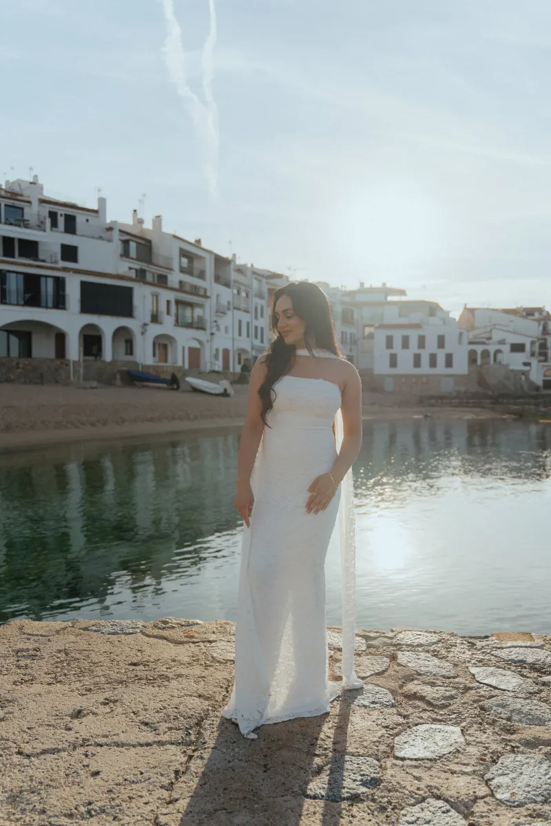 Woman in a white dress smiles by the waterfront at sunset, captured during a surprise proposal photoshoot in a coastal town.