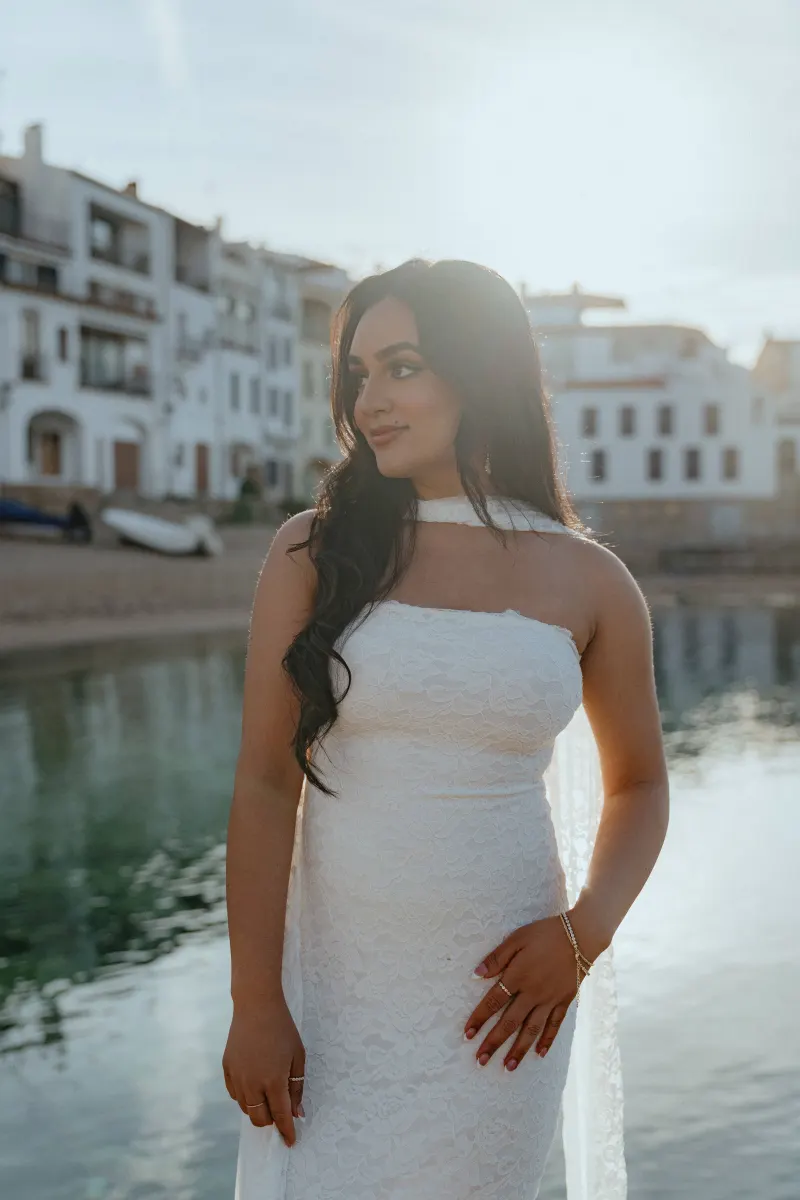 Woman in a white dress smiles by the waterfront at sunset during a surprise proposal photoshoot in a coastal village.
