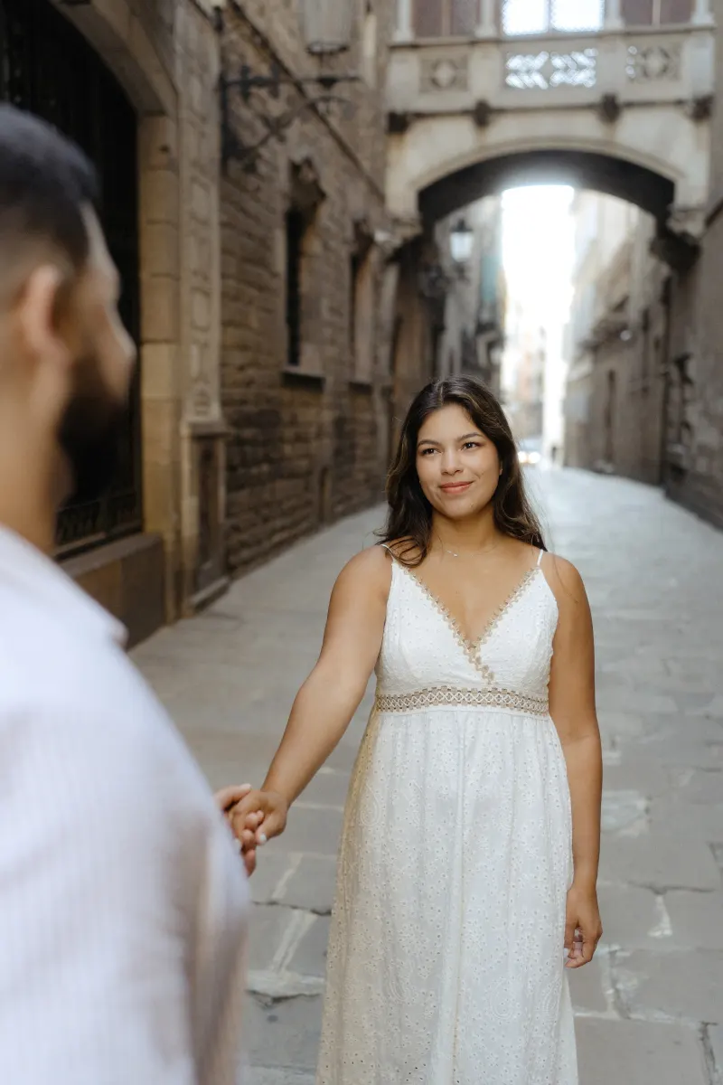 Woman in a white dress smiles, holding her partner’s hand on a quiet historic street during a couples photoshoot.