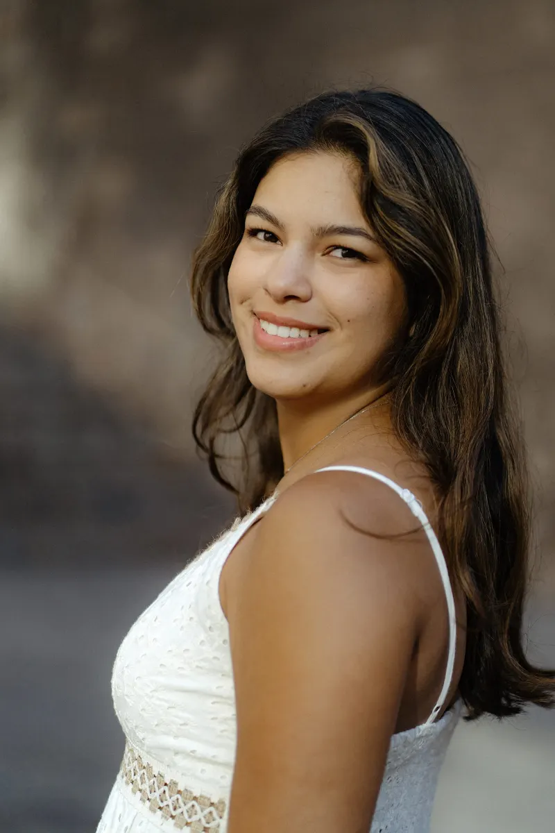 Woman in a white dress smiles softly at the camera, captured during a couples & engagements photoshoot outdoors.