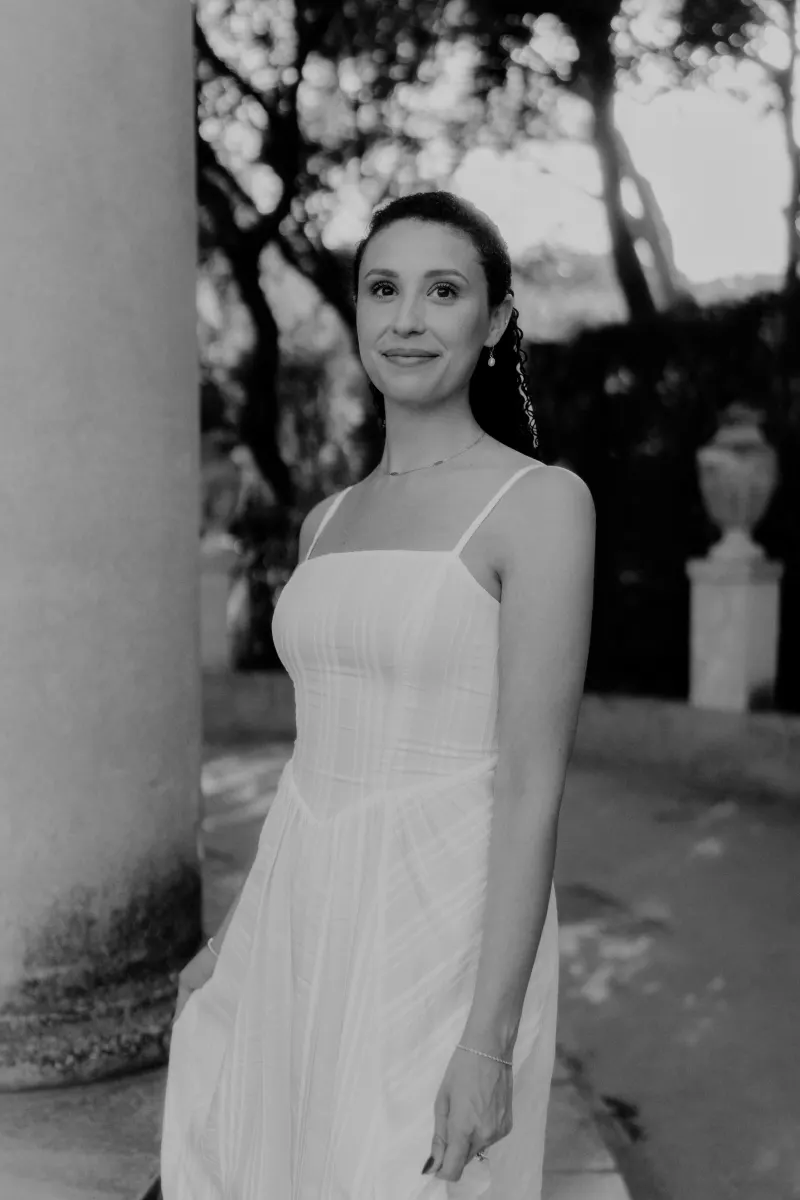 Woman in a white dress smiles softly by a stone column during a couples & engagements photoshoot in a garden setting.