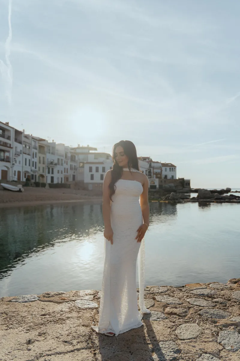 Woman in a white dress smiles softly by the seaside at sunset, captured during a surprise proposal photoshoot.
