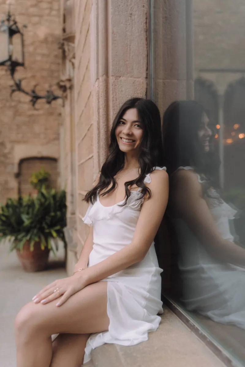 Woman in a white dress smiles softly while seated by a stone wall, captured during a couples & engagements photoshoot.