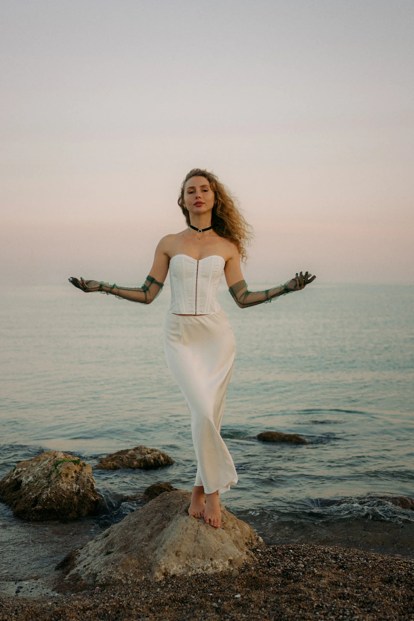 Woman in a white dress stands barefoot on a seaside rock at sunset, serene during a Vacation Portraits photoshoot.