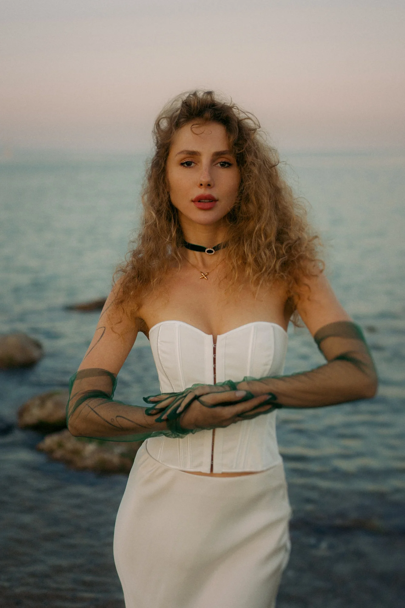 Woman in a white dress stands by the sea at sunset, striking a serene pose during a Vacation Portraits photoshoot.