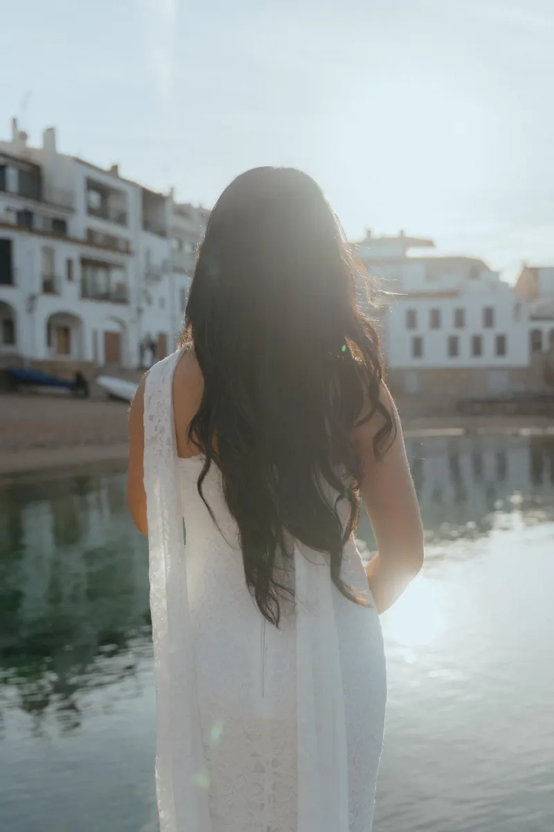 Woman in a white dress stands by the water at sunset, captured during a surprise proposal photoshoot in a coastal village.