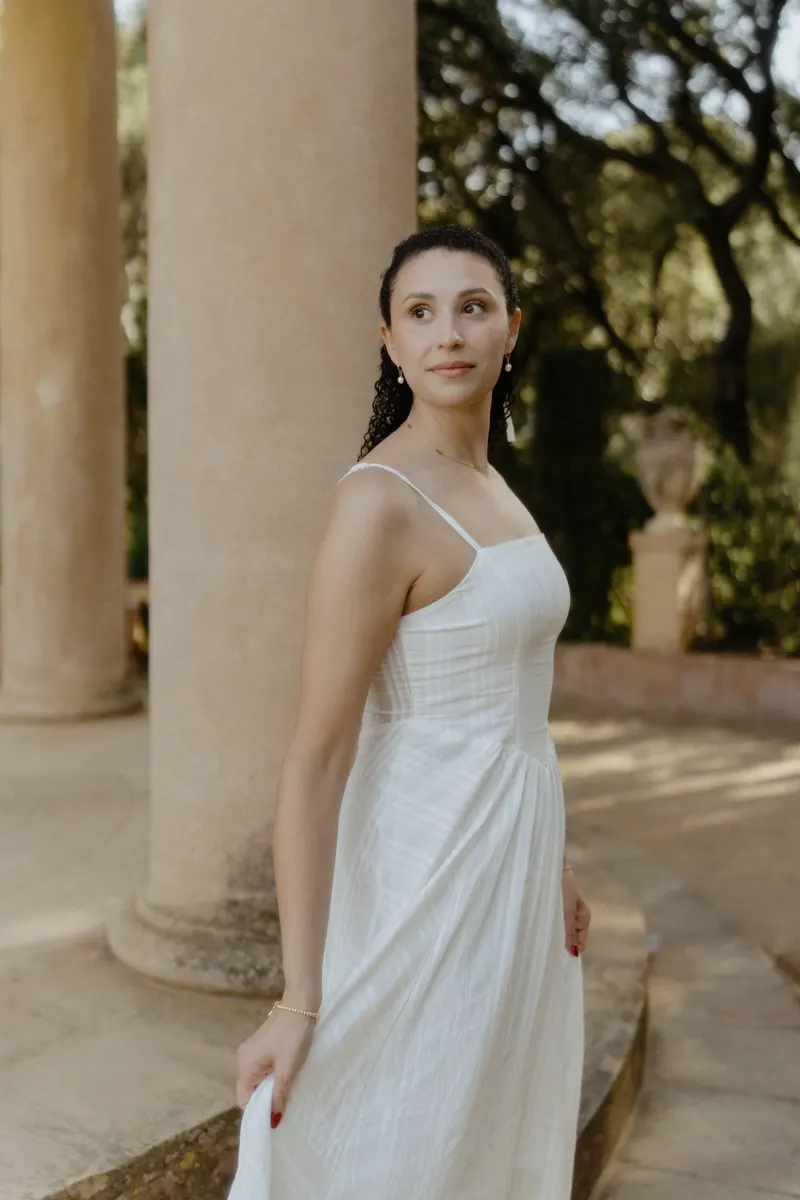 Woman in a white dress stands gracefully by stone columns, captured during a couples & engagements photoshoot.