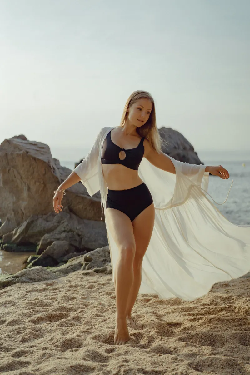 Woman in black swimwear and white cover-up dances barefoot on a sandy beach, captured during a Vacation Portraits photoshoot.