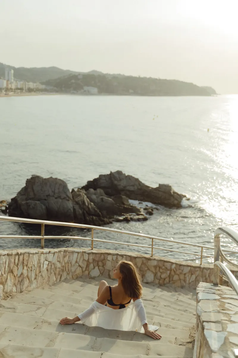 Woman in swimwear sits on seaside steps, gazing at the ocean during a Vacation Portraits photoshoot at sunset.
