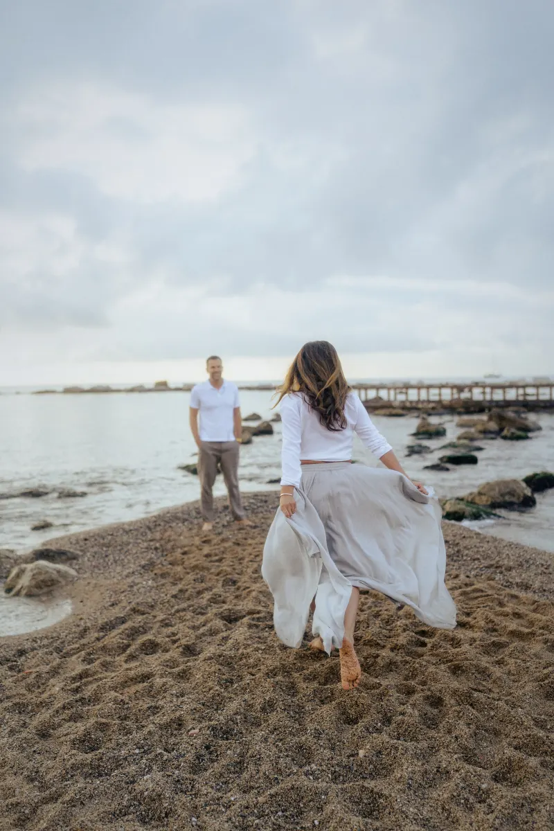 Woman joyfully running barefoot toward her partner on a sandy beach, couples & engagements photoshoot.