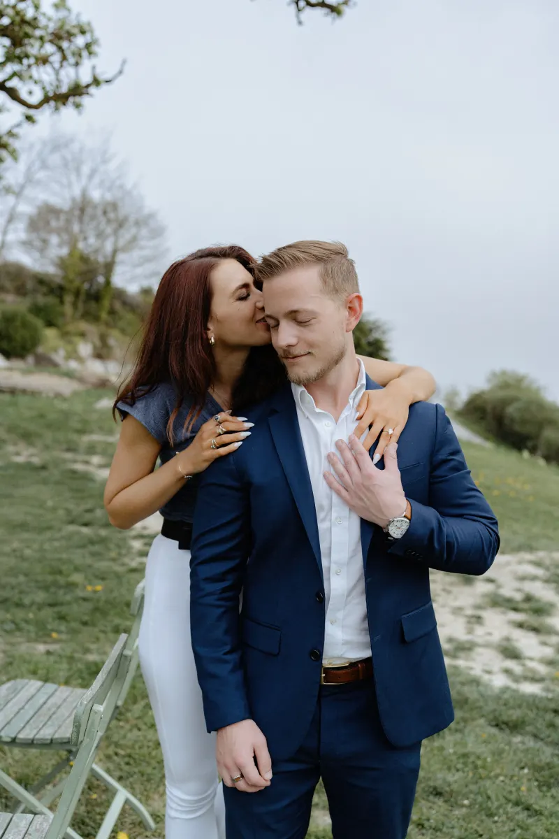 Woman kisses her fiancé’s cheek as they celebrate their engagement during a countryside surprise proposal photoshoot.