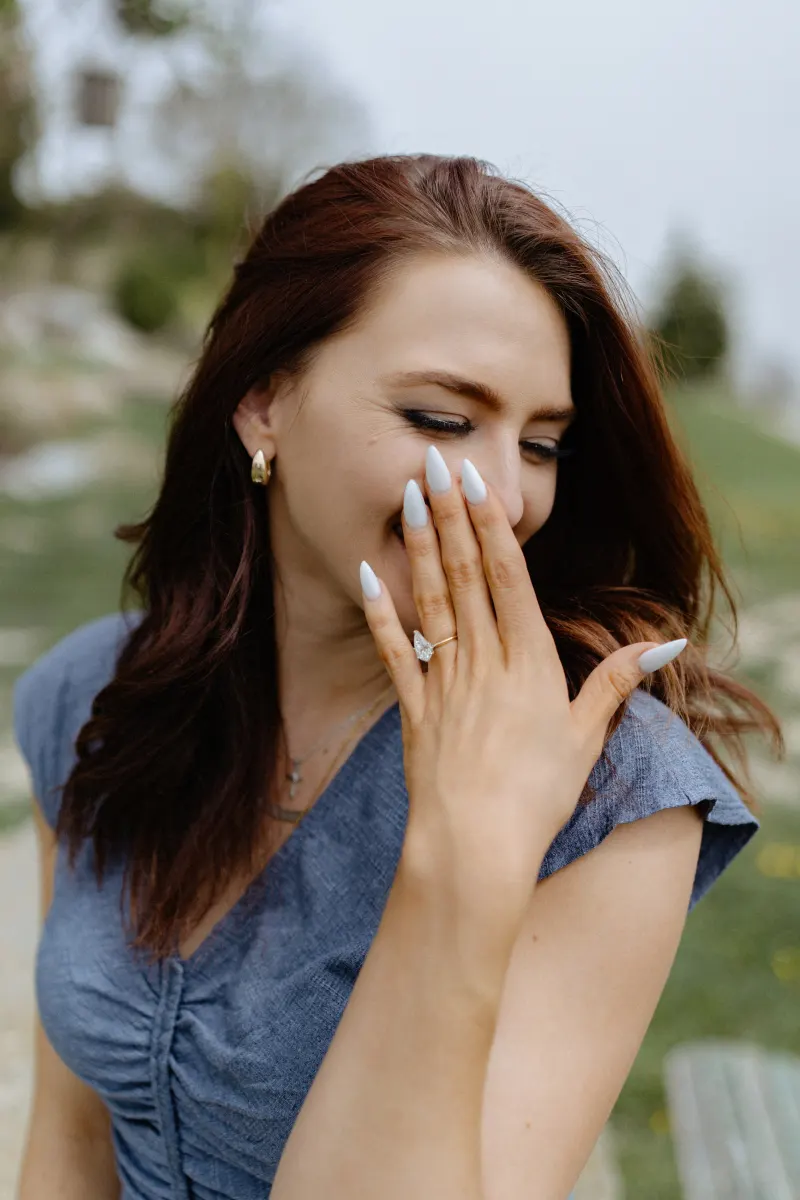 Woman laughs with joy, covering her smile as she admires her engagement ring after a countryside surprise proposal.