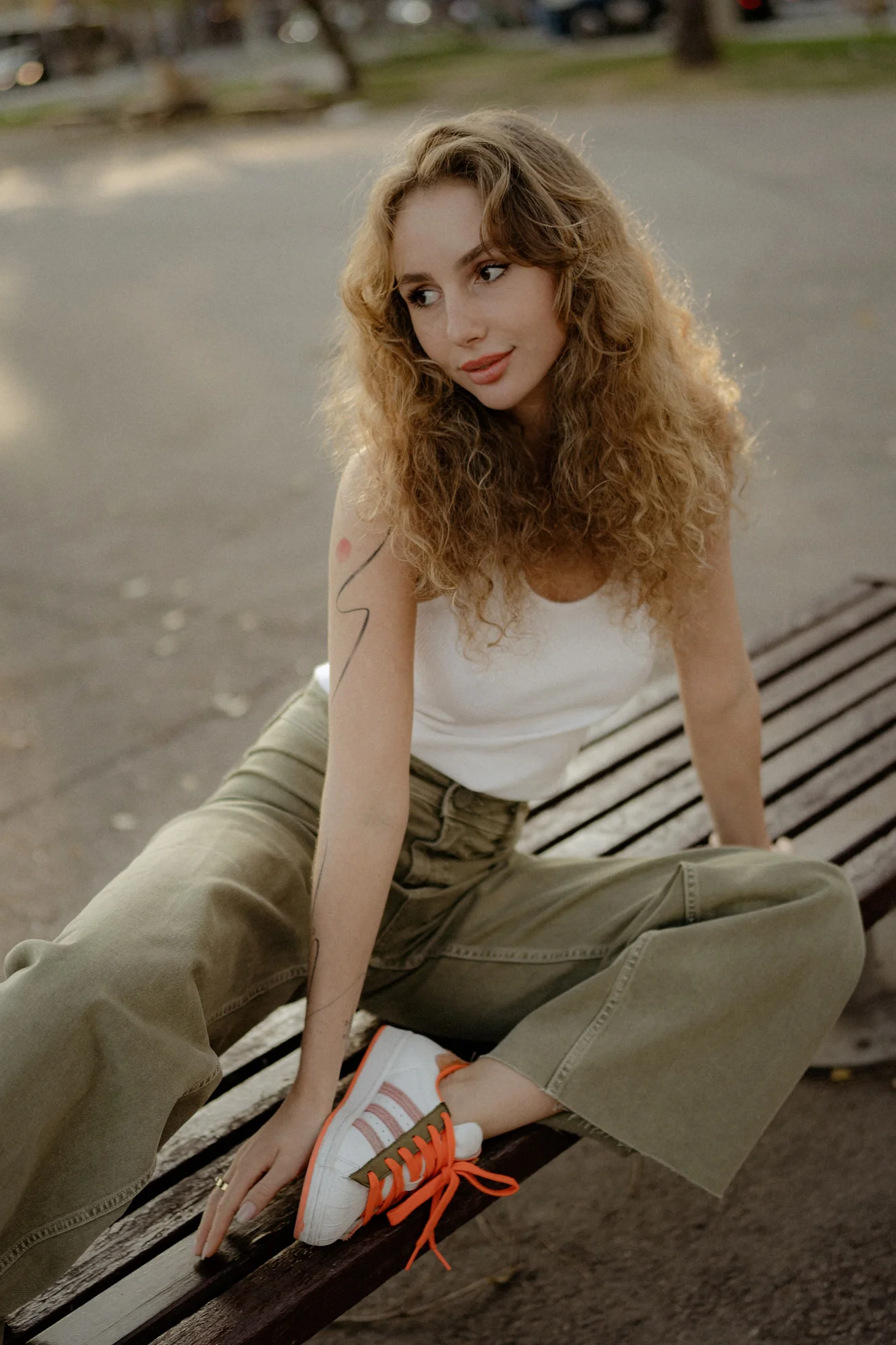 Woman with curly hair sits casually on a park bench, looking away and smiling during a Vacation Portraits photoshoot.