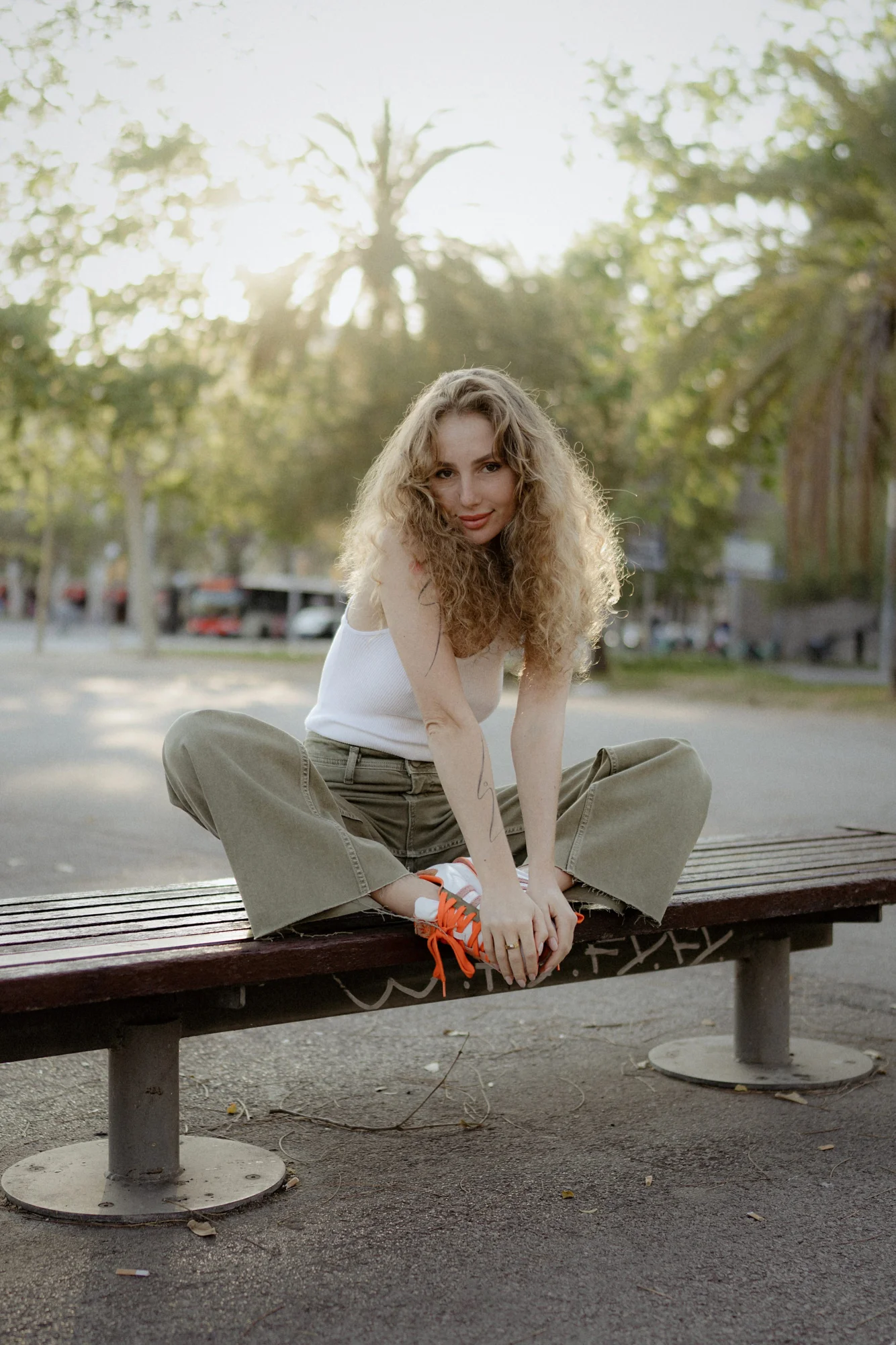 Woman with curly hair sits cross-legged on a park bench, smiling softly during a Vacation Portraits photoshoot.