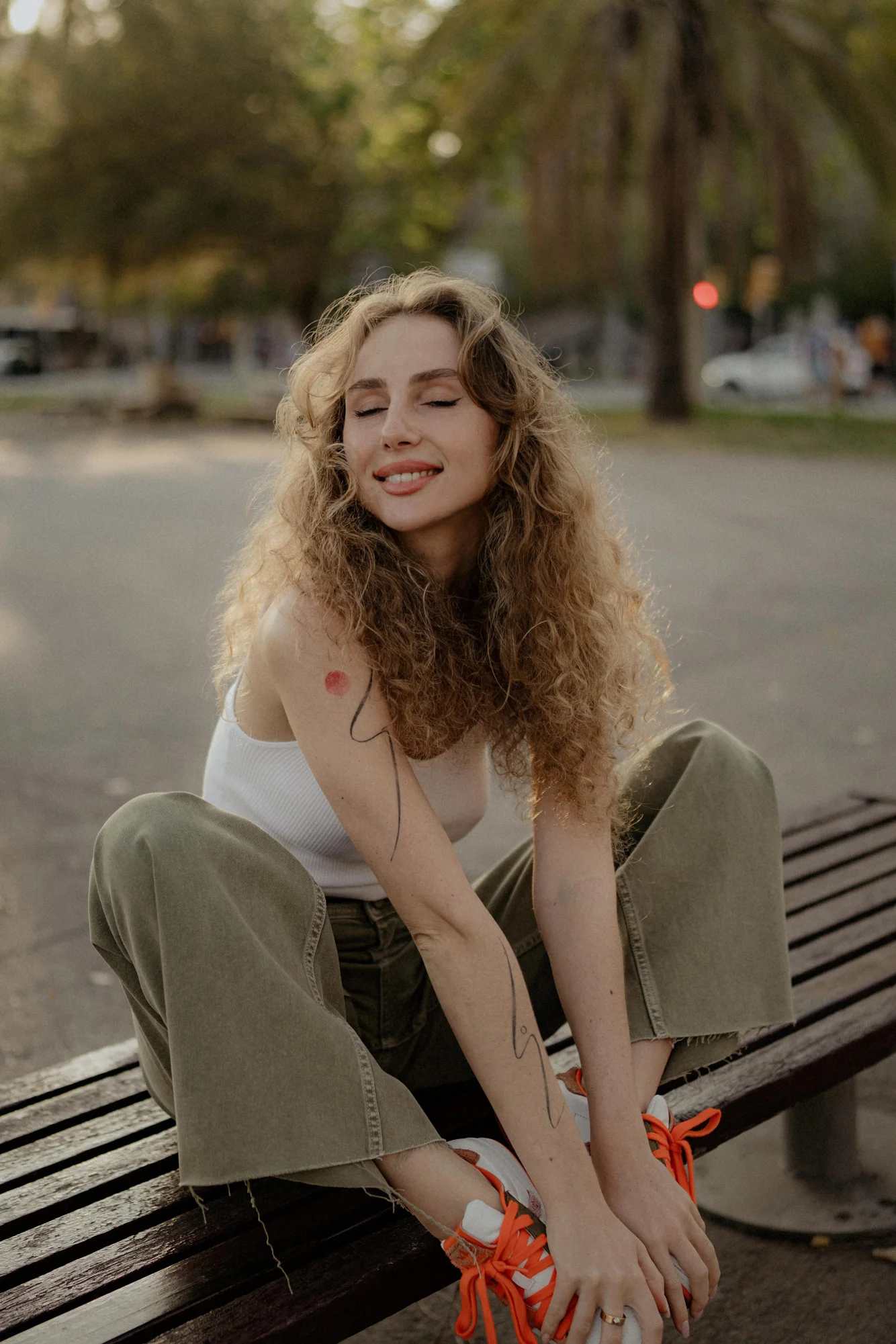 Woman with curly hair smiles with eyes closed, enjoying a relaxed moment on a park bench during a Vacation Portraits shoot.