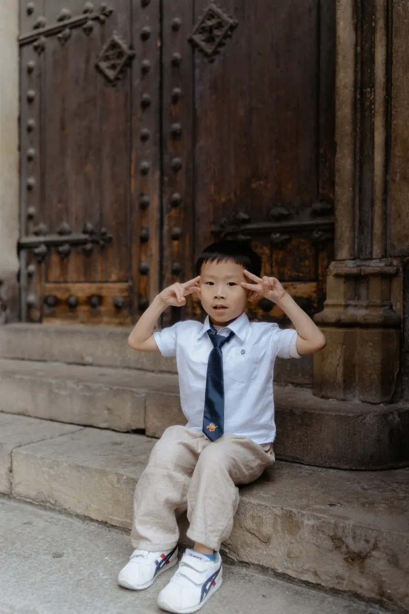 Young boy in dress clothes flashes peace signs while sitting on stone steps during a Vacation Portraits session in Barcelona.