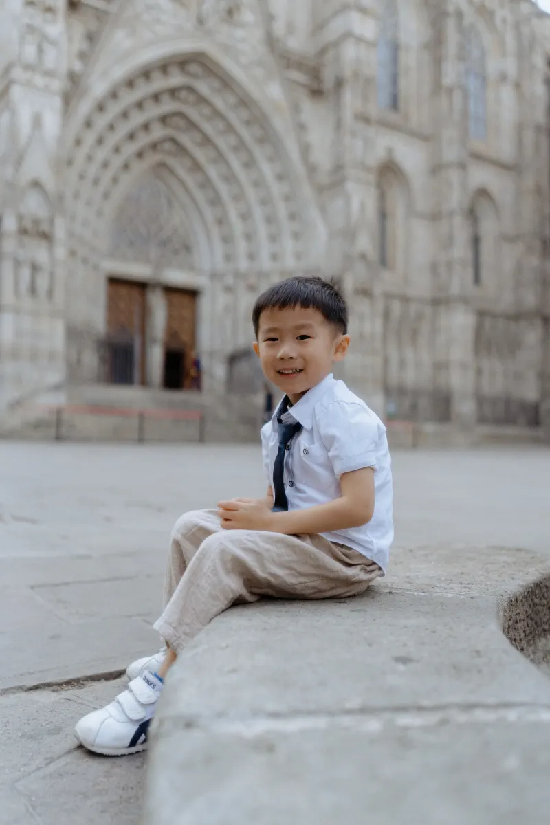 Young boy in dressy clothes smiles while sitting outside Barcelona’s cathedral during a Vacation Portraits photoshoot.