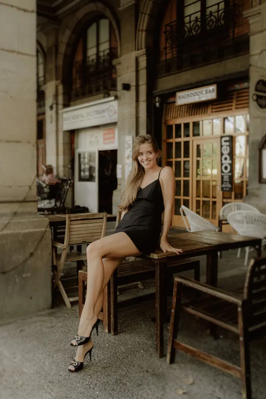 Young woman in a black dress sits on a café table, smiling during a Vacation Portraits session in a European plaza.