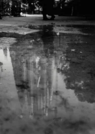 A couple strolls near La Sagrada Familia, its reflection shimmering in a rain puddle, capturing a quiet romantic moment.