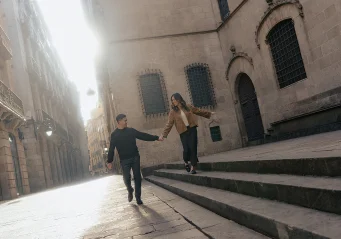 Couple holding hands and laughing as they walk down steps in the sunlit Gothic Quarter, captured by a professional photograph
