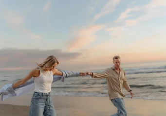 Couple holding hands and laughing while walking along Ginesta beach at sunset, captured during a candid couples session.