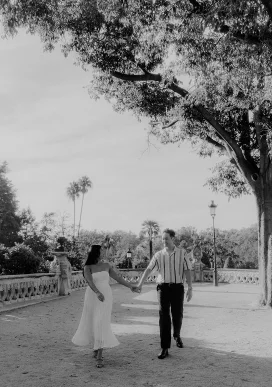 Couple holding hands and smiling while walking under trees in Park de la Ciutadella, captured during a romantic photoshoot.