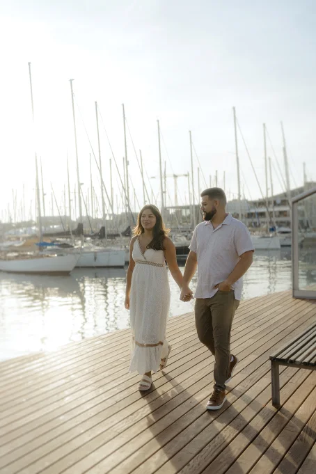 Couple holding hands and strolling along the marina at Port Vell, Barceloneta during a golden hour photo session.