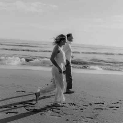 Couple laughing and walking barefoot along Barceloneta beach, candid moment captured by a Barcelona photographer.