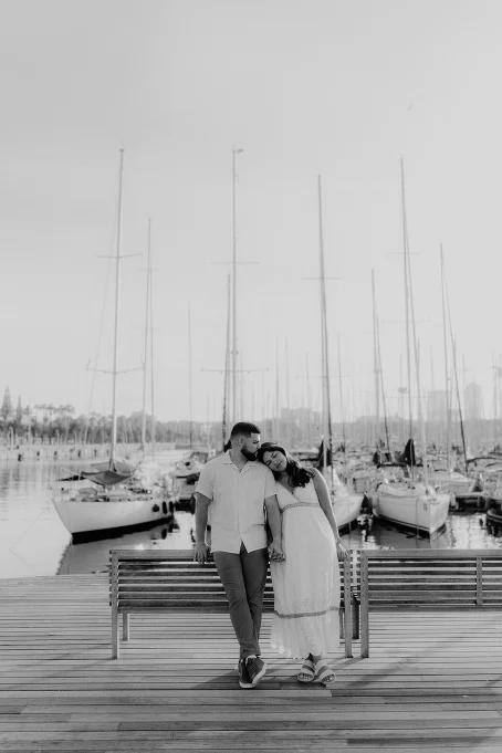 Couple sharing a tender moment on a bench at Port Vell, Barceloneta, with sailboats in the background.