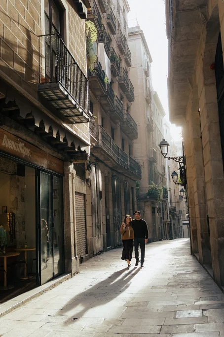 Couple strolling arm in arm through a sunlit street in the Gothic Quarter, captured by a professional photographer.