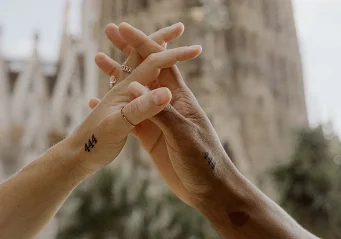 Couple’s hands intertwined, matching tattoos visible, with La Sagrada Familia softly blurred in the background.