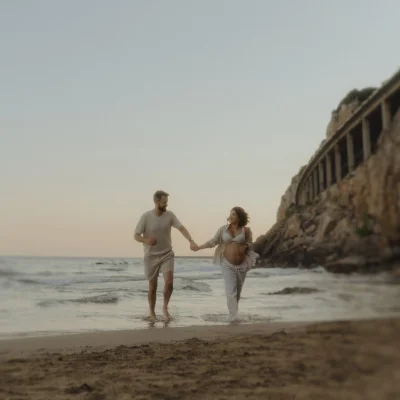Expectant couple holding hands and smiling while walking along Ginesta beach at sunset, maternity photography session.