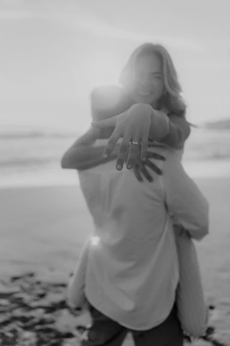 Joyful couple celebrates engagement at Ginesta beach, woman showing ring while embraced at sunset.