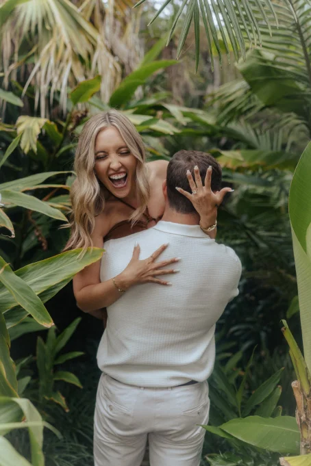 Joyful couple embraces in lush Marimurtra garden, capturing candid engagement photography in a tropical setting.