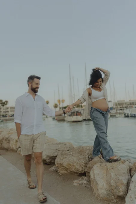 Joyful couple enjoying a maternity photoshoot at Ginesta beach and port, holding hands by the marina.