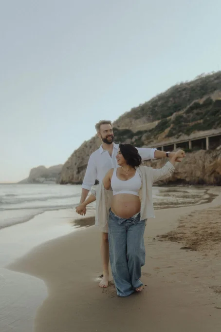 Joyful couple expecting a baby walks hand in hand along Ginesta beach, captured in a relaxed maternity session.