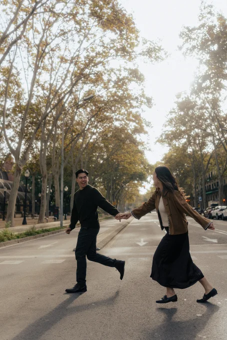 Joyful couple holding hands and laughing while crossing a tree-lined street in the Born area, location photography session.