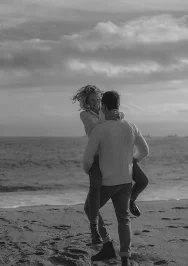 Joyful couple laughing as he lifts her on Barceloneta beach, capturing candid love at Port Vell for a couples photoshoot.
