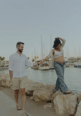 Joyful maternity couple walking hand in hand by the boats at Ginesta beach and port, capturing a tender moment.