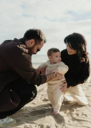 Parents help their baby take first steps on Ginesta beach, capturing a candid family photography moment.